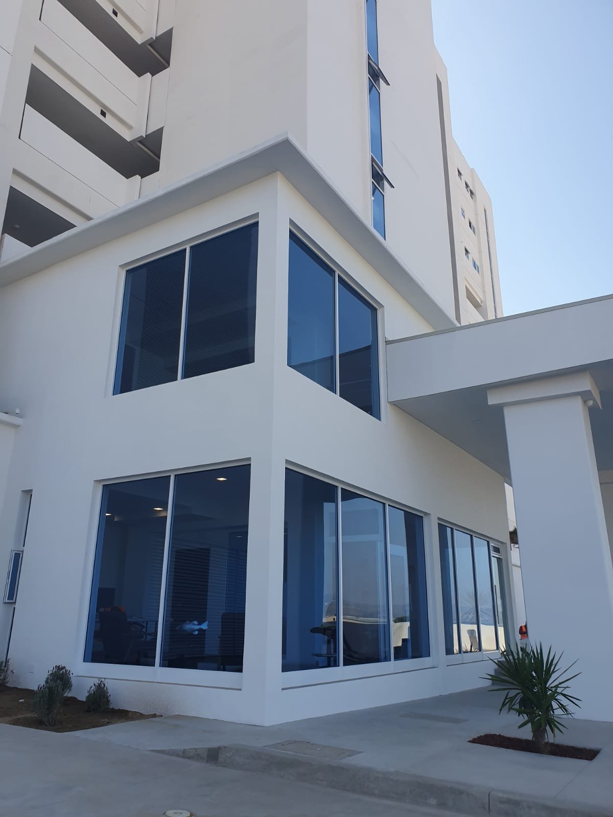 Edificio blanco moderno con ventanas de marco oscuro, torre residencial de varios pisos al fondo, cielo azul claro