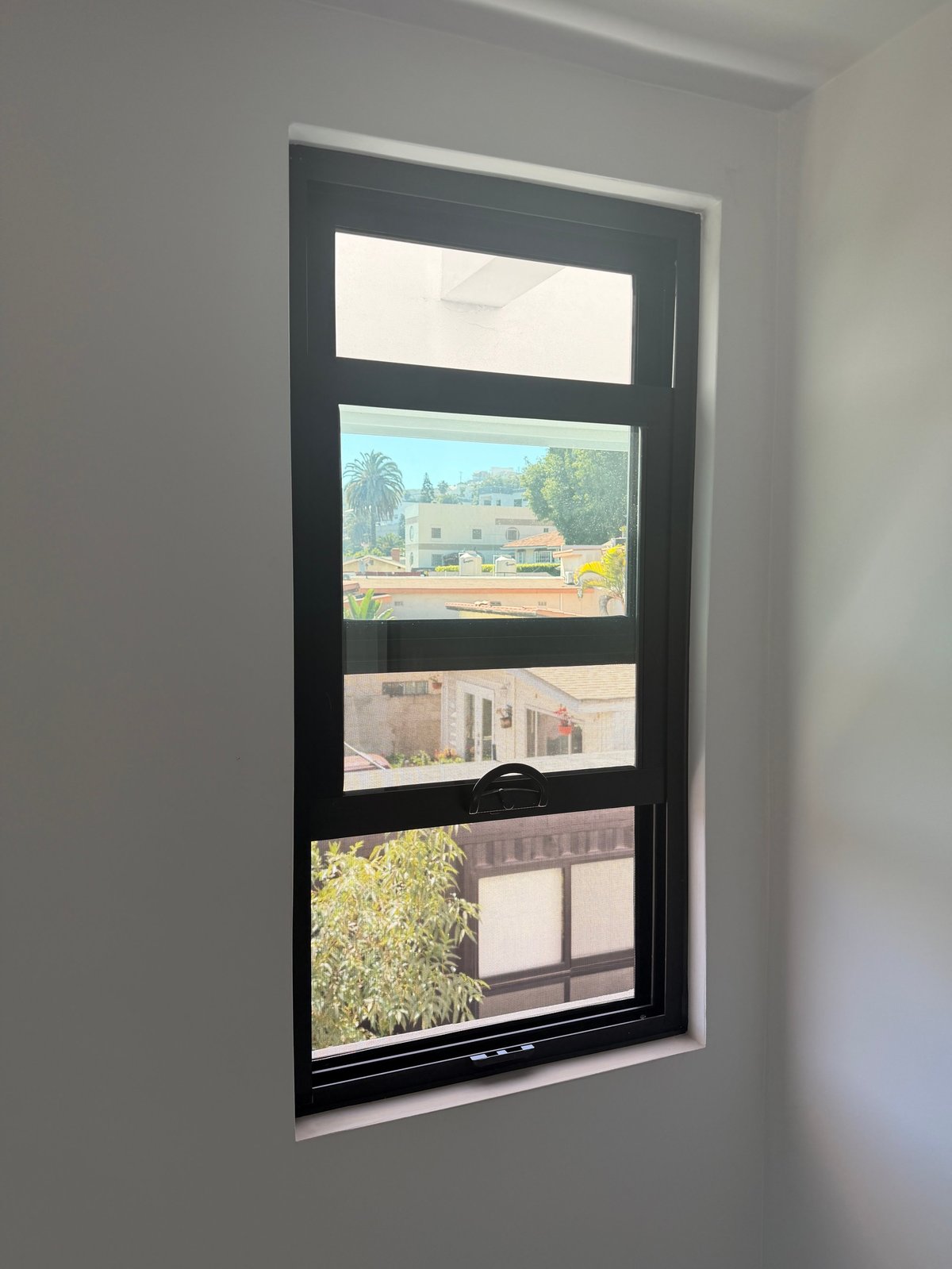 Ventana de cuatro paneles con marco negro montada en pared blanca, mostrando vistas de edificios, palmeras y jardinería exterior