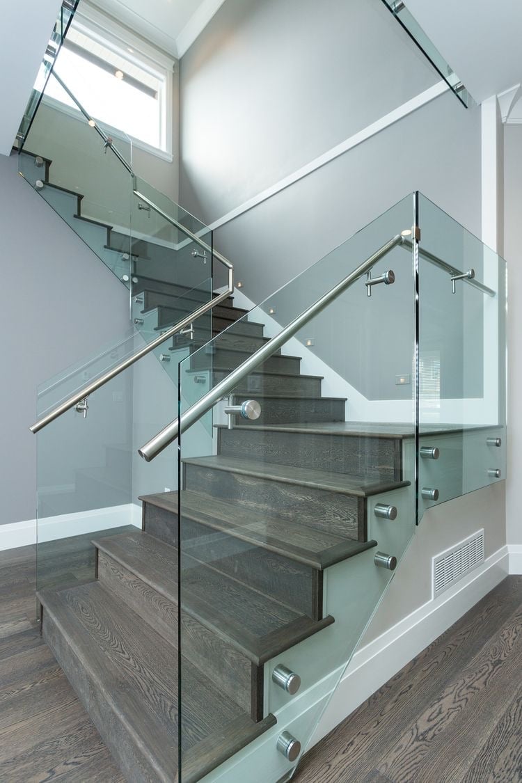 Escalera moderna con escalones de madera oscura, barandales de vidrio, pasamanos de metal y tragaluces en espacio interior contemporáneo brillante