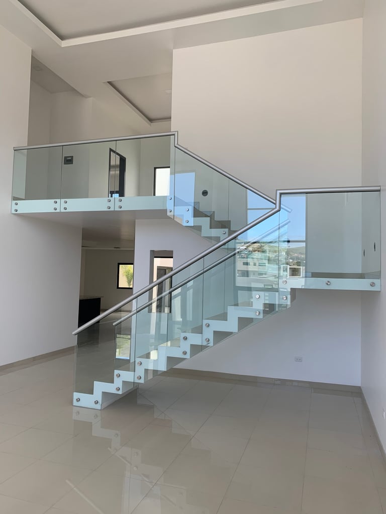 Escalera moderna de vidrio con barandal de metal en espacio interior vacío con paredes blancas y piso de loseta