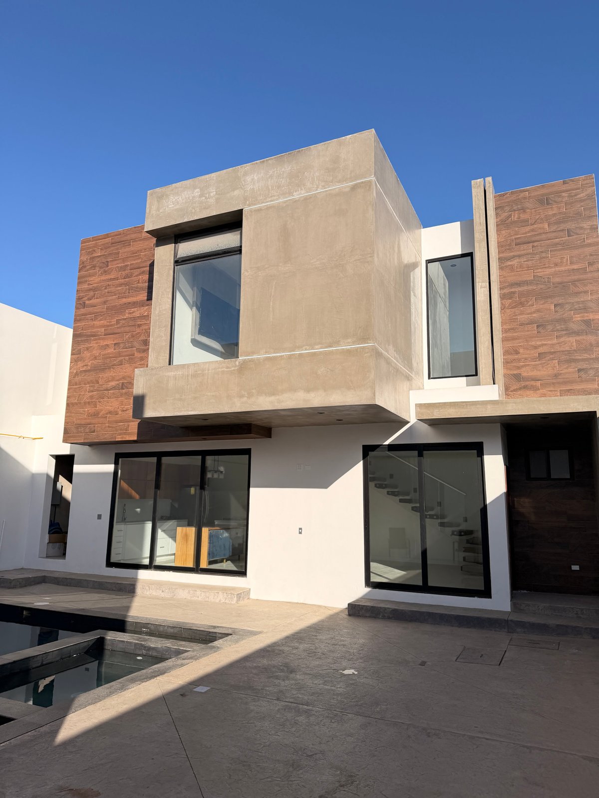 Casa moderna de dos pisos con diseño geométrico, concreto beige, ladrillo rojo, acabado blanco y grandes ventanas contra cielo azul